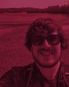 a heavily tinted photo of a mad smiling at the camera. he has thick wavy hair and a beard and is wearing sunglasses